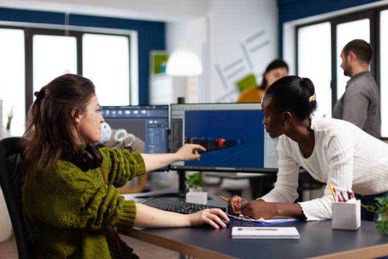 Diverse women game developers creating game interface sitting in startup creative production company pointing at pc displays. Workers developing online graphics video games with modern technology